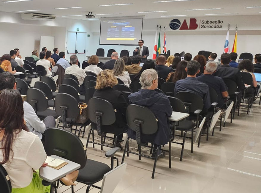 auditório da OAB Sorocaba visto do fundo da sala, palestrante ao fundo junto ao púlpito, primeiras fileiras da plateia em primeiro plano levemente desfocadas. Luz de ambiente real, sem cara de banco de imagens.
Alt text: Palestra sobre reforma tributária no planejamento imobiliário na OAB Sorocaba