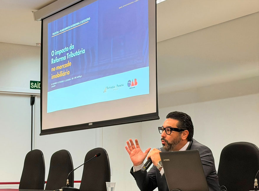mãos do palestrante em gesto de explicação, tela de projeção ao fundo com as palavras "holding" e "permuta" legíveis; rosto cortado pelo enquadramento.
Alt text: Debate sobre holdings e permutas diante da reforma tributária imobiliária