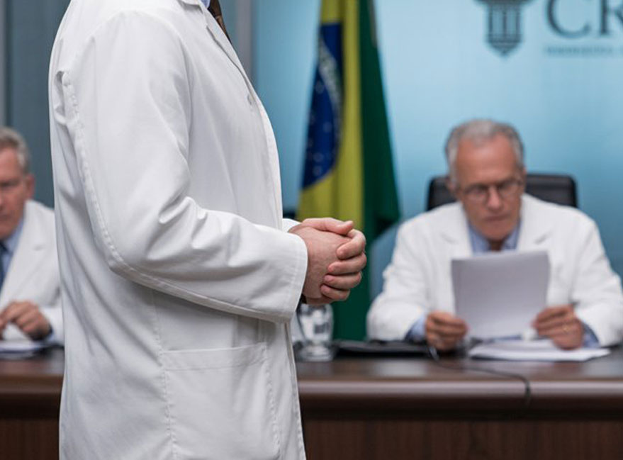 Um médico de jaleco (rosto fora do quadro) sentado a uma mesa, revisando com atenção uma pasta de documentos; ao lado, as mãos de uma advogada apontando um trecho do papel. Sobre a mesa, uma caneta, um carimbo e uma pasta com etiqueta neutra. Luz natural de janela, profundidade de campo curta — o documento em foco, o ambiente desfocado. Ao fundo, muito borrado, um consultório sóbrio. Paleta neutra e clínica (branco, cinza, azul discreto). Estética jornalística, sem rostos nítidos, sem texto legível, sem logotipos, sem aparência de banco de imagens.